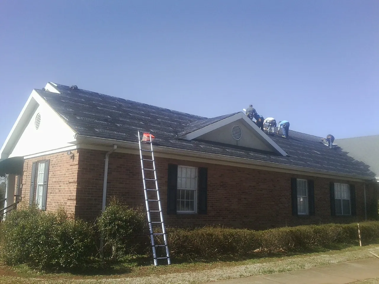 Crew tearing shingles off a brick ranch roof with a ladder set — Emergency Roof Repair work by our crew in Schoenchen