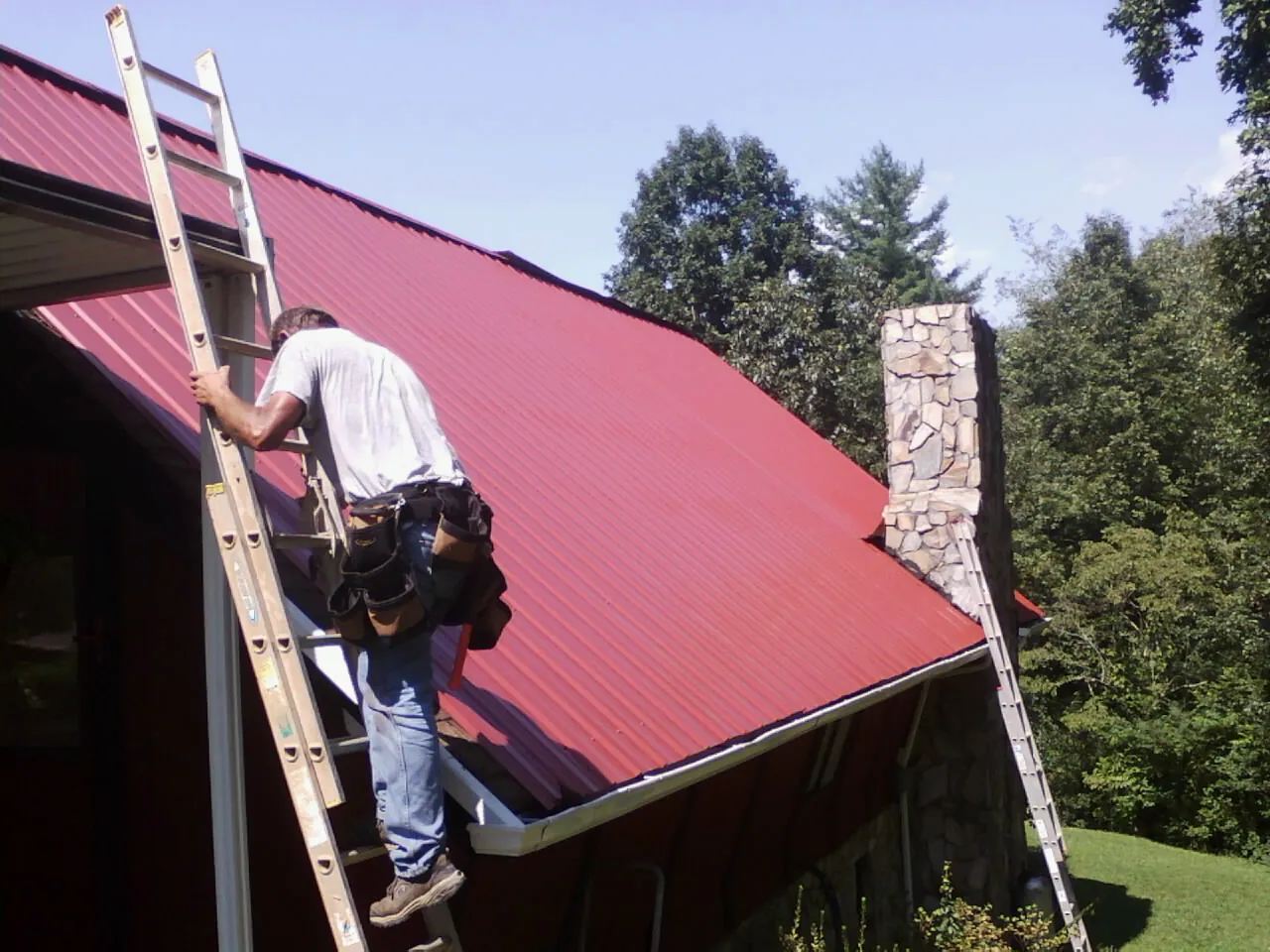 Roofer on a ladder beside a red metal roof and stone chimney — Soffit & Fascia Repair by Roofing Brothers in Schoenchen